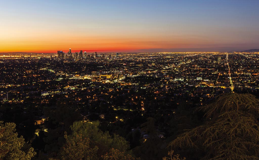 A view of city lights from a hill. The sunset light glows red-orange below a dim blue sky.