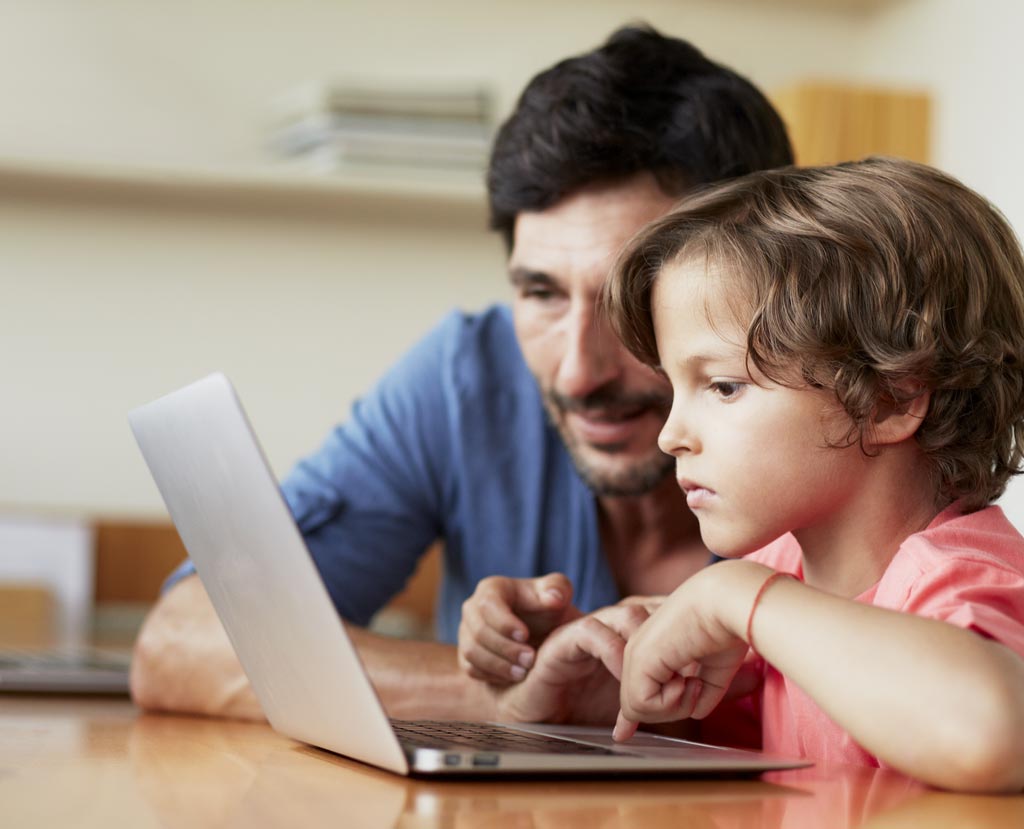 A man with dark hair and light-colored skin sits next to a boy with similar complexion, showing him something on a laptop.