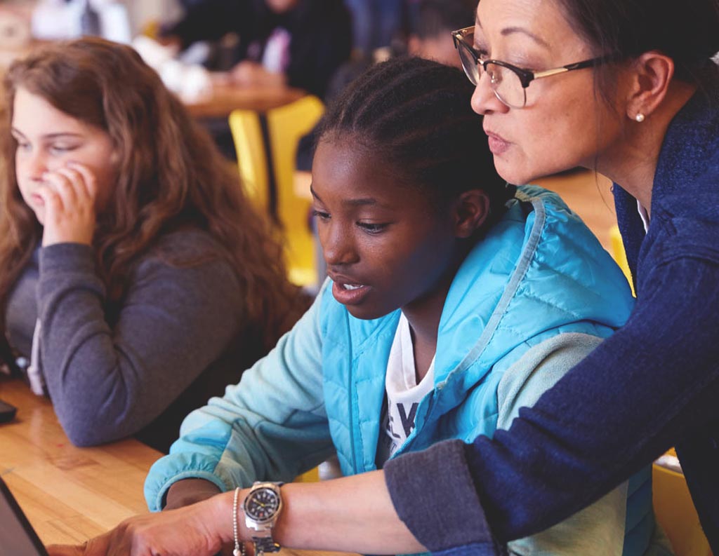 An Asian female teacher with glasses sits behind a Black female student in a classroom, pointing to something off-screen.